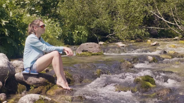Tourist-woman-sitting-on-shore-of-stony-river-and-resting-while-summer-hike