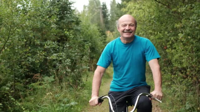 Senior-hombre-caucásico-en-camiseta-azul-disfrutando-de-sus-vacaciones-de-verano,-andar-en-bicicleta-al-aire-libre-entre-los-árboles.