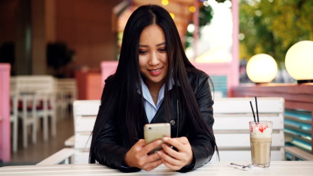 Lächelnde-asiatische-Studentin-ist-ruhen-im-Café-im-freien-und-mit-Smartphone-Bildschirm-zu-berühren-und-zu-beobachten.-Social-Media,-moderne-Technologie-und-City-Konzept.