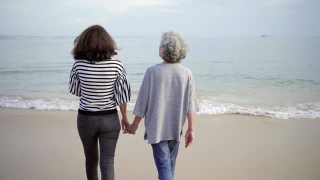 Back-view-of-two-happy-mature-ladies-holding-hands-together-while-walking-on-sandy-beach.