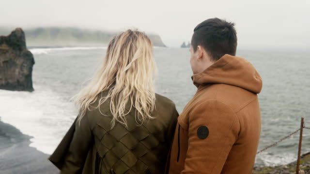 Jóvenes-viajar-pareja-en-playa-negra-en-Islandia-buscando-en-los-pies-de-troll-rocas,-famoso-vista,-sintiéndose-feliz,-levanta-la-mano