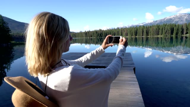 Young-woman-standing-on-wooden-pier-above-lake-takes-a-photo-of-stunning-mountain-lake-scenery-using-her-mobile-phone