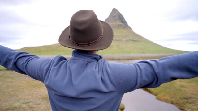 4K---junger-Mann-in-Island-Arme-ausgestreckt-für-Freiheit-Frühling-bedeckten-Himmel-am-berühmten-Berg-Kirkjufell