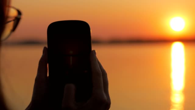 woman-is-taking-photo-of-a-beautiful-vivid-sunset-over-river,-using-her-smartphone,-close-up