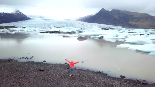 DRONE-mit-Blick-auf-Frauen-stehen-durch-Gletscherlagune-in-Island-ausgestreckten-Armen