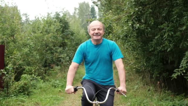 Senior-caucasian-male-in-blue-t-shirt-enjoying-his-summer-vacation-riding-a-bicycle-outdoor-between-trees.