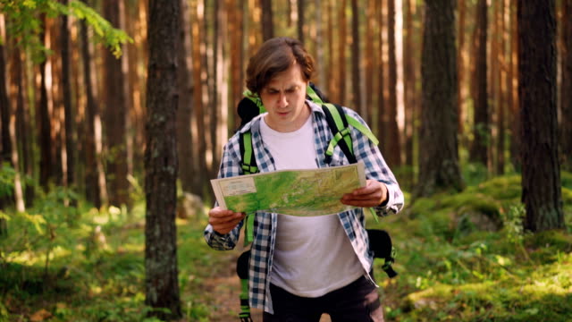 Smiling-young-man-is-looking-at-map-and-searching-for-right-way-in-forest-while-multiethnic-group-of-friends-is-following-him.-Friendship,-nature-and-people-concept.