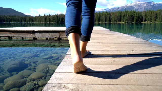 Cerrado-los-pies-de-la-mujer-caminando-en-el-muelle-de-madera-sobre-el-impresionante-paisaje-de-lago-de-montaña
