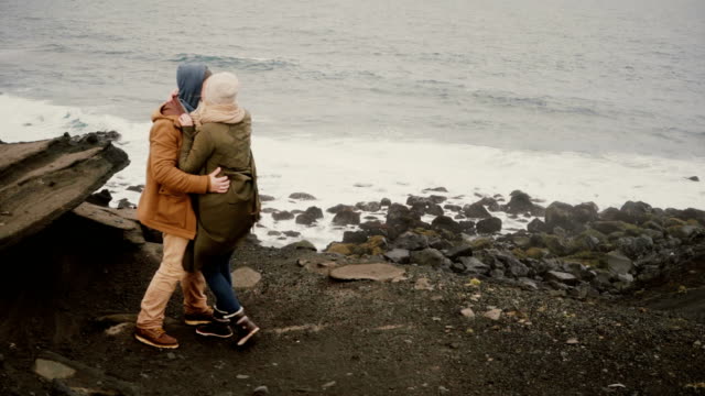 Back-view-of-young-man-and-woman-standing-on-the-shore-of-the-sea-in-cold-day,-holding-hands-and-looking-on-water