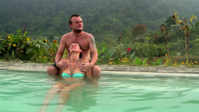 Woman-relaxes-in-a-hot-spring-with-thermal-water.-Man-sits-on-the-edge-of-the-pool-and-hugs-a-woman