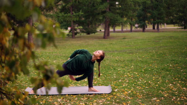 Yoga-student-is-practising-variations-of-Side-Angle-position-Parsvakonasana-on-yoga-mat-in-park.-Green-and-yellow-trees,-leaves-and-grass-are-visible.