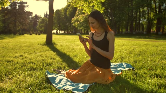 Young-lady-enjoying-Nature-and-sunset.-Sitting-on-green-summer-park-using-mobile