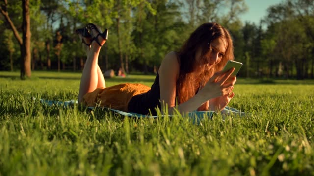 Happy-female-types-sms-or-chat,-sitting-in-park-on-grass.-Woman-chatting-in-telephone