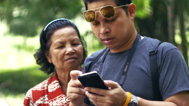 Asiatische-Mutter-und-Sohn-im-Garten-während-Sohn-mit-Smartphone-Morgen-ruhen.