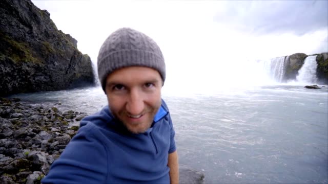 Junger-Mann-unter-Selfie-Porträt-mit-herrlichen-Wasserfall-in-Island,-Godafoss-fällt.-Die-Leute-reisen-Exploration-Konzept