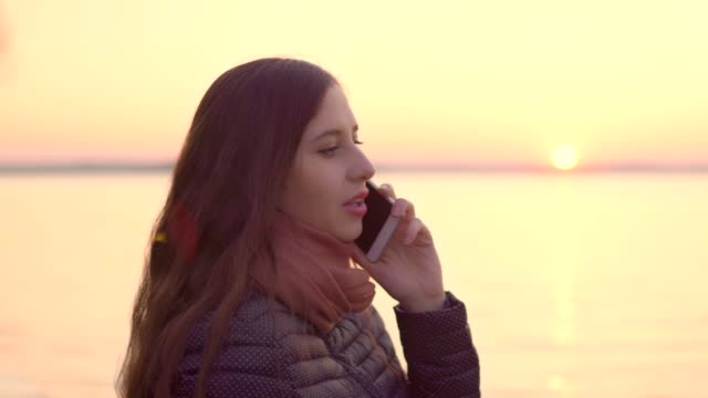 Mujer-atractiva-joven-hablando-por-teléfono-móvil-en-el-fondo-puesta-de-sol-en-el-lago.-4k