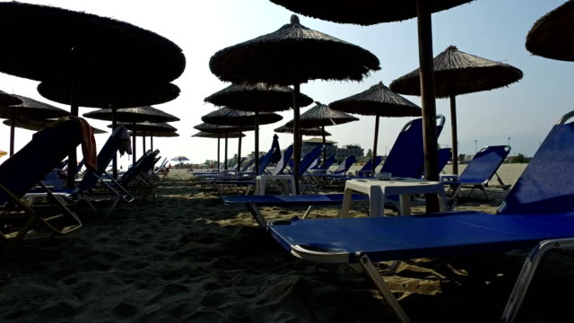 A-pie-de-playa-de-arena-entre-sombrillas-y-camas-lounge.-Vacaciones-en-el-mar,-playa-y-sombrillas-de-paja-bajo-el-cielo-azul,-4-k-tiro-de-steadicam