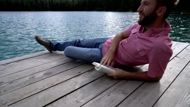 Young-man-relaxing-on-lake-pier-with-book,-Canada