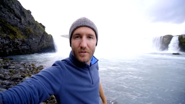Junger-Mann-unter-Selfie-Porträt-mit-herrlichen-Wasserfall-in-Island,-Godafoss-fällt.-Die-Leute-reisen-Exploration-Konzept