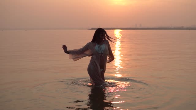 Sexy-girl-in-a-beach-tunic-dancing-standing-in-the-water-against-a-sunset-background.-slow-motion