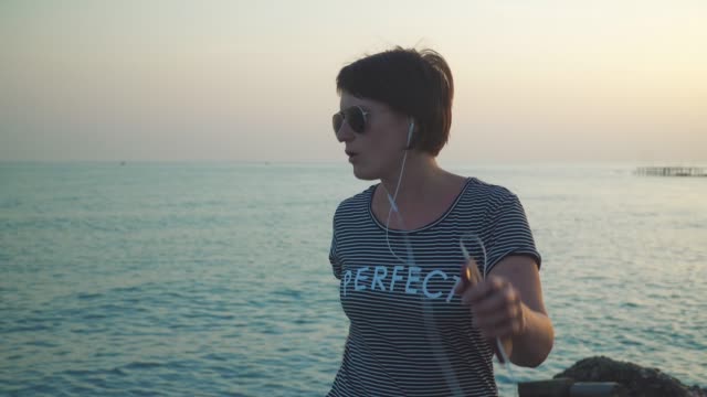Mujer-camiseta-escuchando-música-en-auriculares-y-bailando-en-ocio-contra-Marina-puesta-del-sol