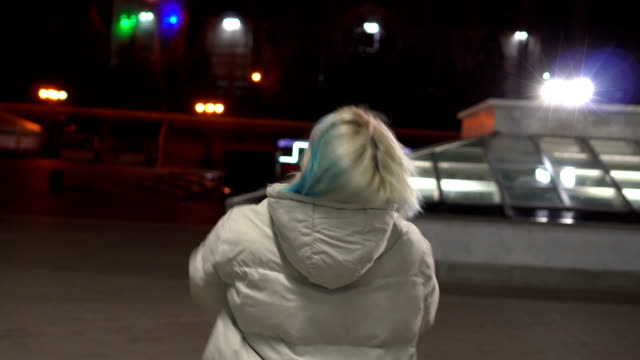 Young-blonde-woman,-in-a-white-fashion-jacket-walking-down-the-deserted-central-street-at-night