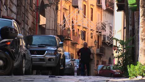 A-man-uses-a-cane-to-walk-down-a-street-pass-cars-lined-against-the-sides-of-buildings-Palermo-Italy