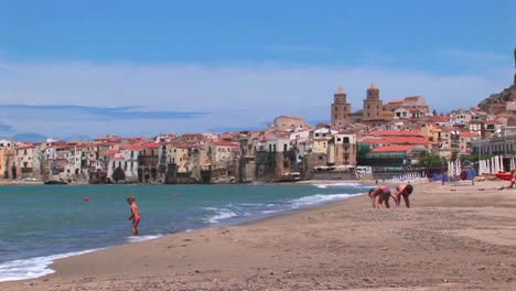 Small-waves-break-near-houses-along-a-shoreline-as-children-play-in-the-sand-in-Cefalu-Italy--