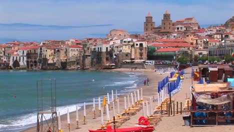 A-small-boat-and-umbrellas-rest-on-a-beach-with-houses-near-the-shoreline-in-Cefalu-Italy----