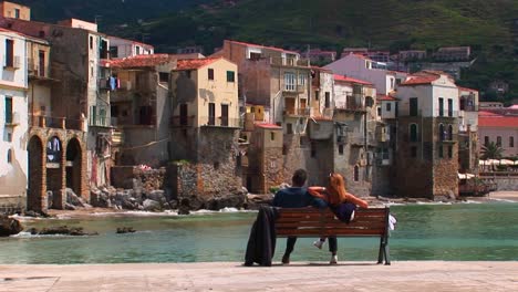 A-couple-sits-on-a-bench-overlooking-the-ocean-and-houses-in-Cefalu-Italy----