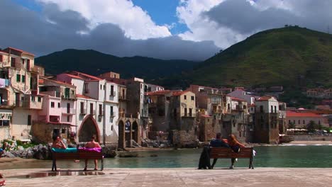 -couples-sit-on-a-benches-overlooking-the-ocean-and-houses-in-Cefalu-Italy-----
