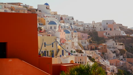 Casas-Multicolores-Bordean-Las-Laderas-De-La-Isla-Griega-De-Santorini-Con-Una-Bandera-Griega-En-La-Distancia