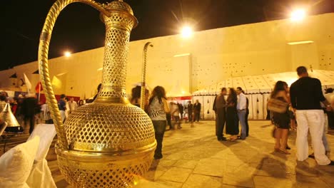 Un-Lapso-De-Tiempo-De-Personas-Caminando-Pasan-Un-Recipiente-De-Líquido-De-Color-Dorado-Por-La-Noche-En-Marruecos