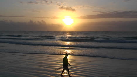 A-man-walks-along-the-beach-carrying-a-stick-as-the-waves-come-in-and-the-sun-sets