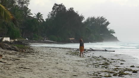 A-woman-walks-along-a-beach-waving-her-arms-a-dog-runs-along-and-the-waves-roll-in