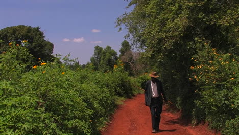 A-man-carrying-a-stick-walks-through-a-grove-of-trees-and-bushes-some-with-orange-flowers