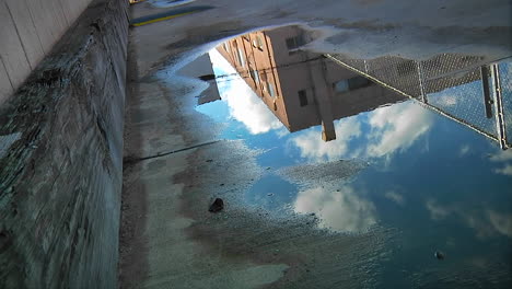 Lapso-De-Tiempo-De-Nubes-Pasando-Sobre-Un-Edificio-Urbano-Como-Se-Refleja-En-Un-Charco-En-La-Acera