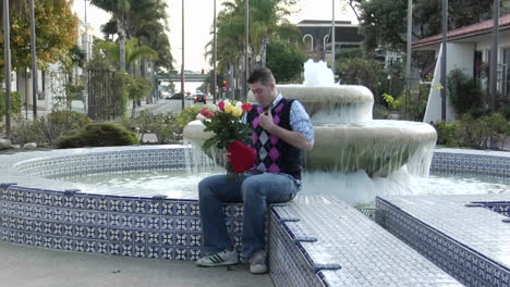 Ein-Mann-Wartet-Aufgeregt-An-Einem-Brunnen-Mit-Blumen-Und-Einem-Herzförmigen-Kasten