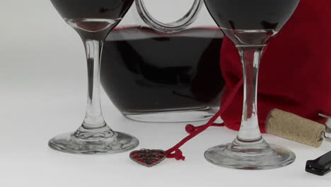 Mit-Rotwein-Gefüllte-Gläser,-Umgeben-Von-Einer-Halbvollen-Karaffe-Und-Rotem-Samtbeutel,-Werden-Auf-Einer-Weißen-Fläche-Präsentiert