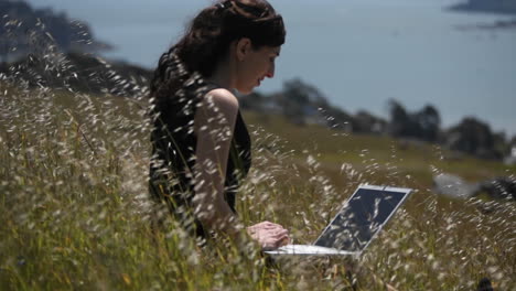 A-woman-using-a-laptop-sits-in-a-field-by-the-shore