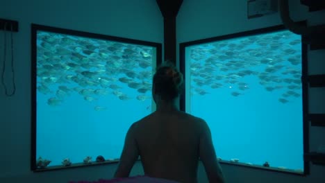 Shirtless-Man-Stands-In-Underwater-Hotel-Looking-At-Fish-Swimming-By-From-His-Window-Tanzania-Africa