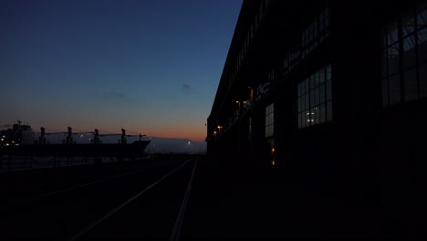 Wide-shot-of-a-large-warehouse-or-factory-at-dusk-or-sunset-as-a-large-cargo-ship-passes-in-the-distance