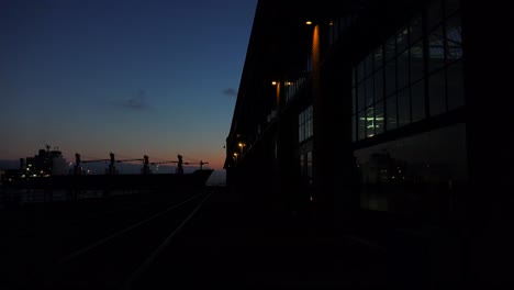 Wide-shot-of-a-large-warehouse-or-factory-at-dusk-or-sunset-as-a-large-cargo-ship-passes-in-the-distance-1