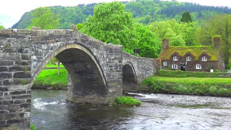 Un-Hermoso-Puente-Sobre-Un-Río-Con-Una-Casa-Cubierta-De-Musgo-En-La-Distancia-Gales