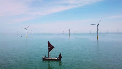 A-flat-bottomed-sailing-barge-sailboat-moves-up-the-Thames-River-Estuary-in-England-amidst-numerous-wind-turbine-windmills