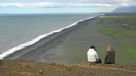 Two-women-sit-on-a-cliff-overlooking-Dyrh_____‚laey-black-sand-beach-Iceland