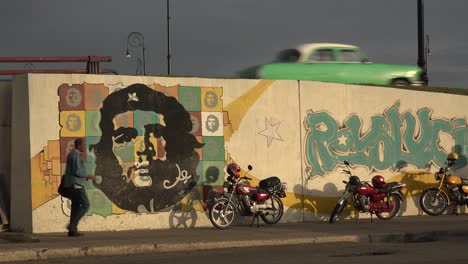 Ein-Auto-Fährt-Eine-Straße-In-Der-Nähe-Eines-Gebäudes-Entlang,-Das-Mit-Revolutionären-Gemälden-Im-Kommunistischen-Kuba-Verziert-Ist