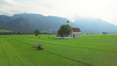 Eine-Schöne-Antenne-über-Einer-Traditionellen-Deutschen-Bayerischen-Kirche-Mit-Einem-Traktor,-Der-Felder-Pflügt