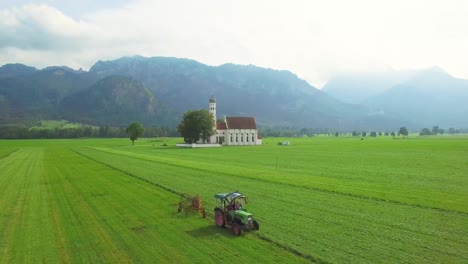 Eine-Schöne-Antenne-über-Einer-Traditionellen-Deutschen-Bayerischen-Kirche-Mit-Einem-Traktor,-Der-Felder-Im-Vordergrund-Pflügt-1