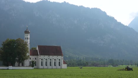 Gründungsaufnahme-Einer-Traditionellen-Deutschen-Bayerischen-Kirche-Mit-Einem-Traktor,-Der-Felder-Im-Vordergrund-Pflügt
